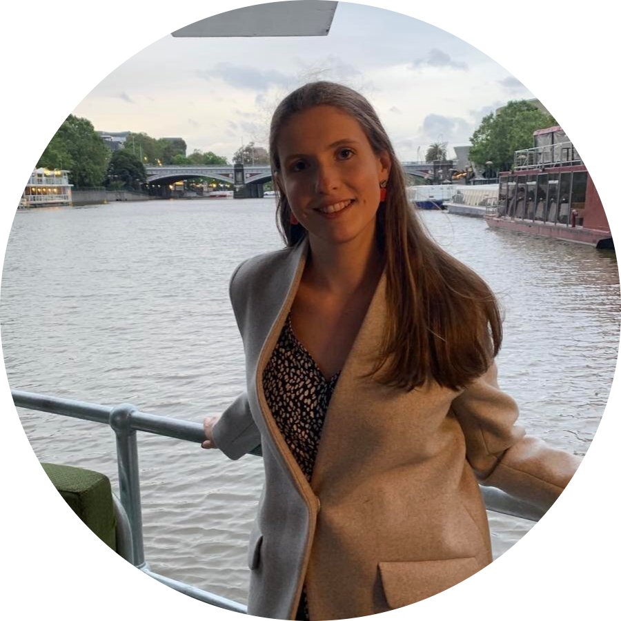 An image of our current secretary, Anna Tinney. Anna is standing against a railing in front of a river background. She's smiling into the camera, and is wearing a smart grey jacket over a black-and-white dress. 