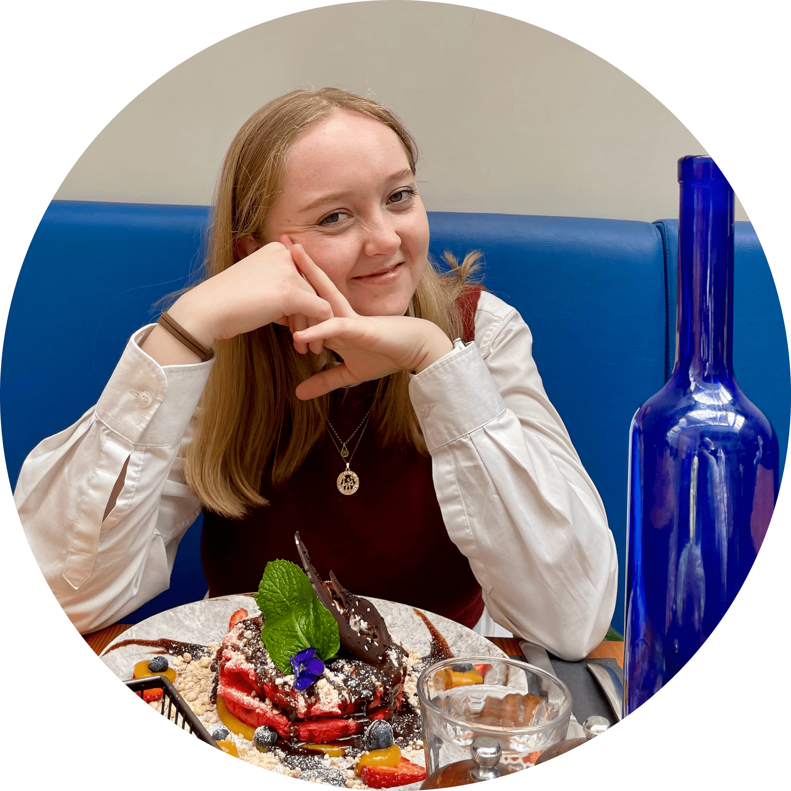 An image of our current treasurer, Jessica Bowen. Jess is smiling with her head propped up by her hands, and is sitting on a blue cushioned booth in front of a fancy-looking plate of sweet food. A drinking glass and blue glass bottle are also visible to the right of the image. 