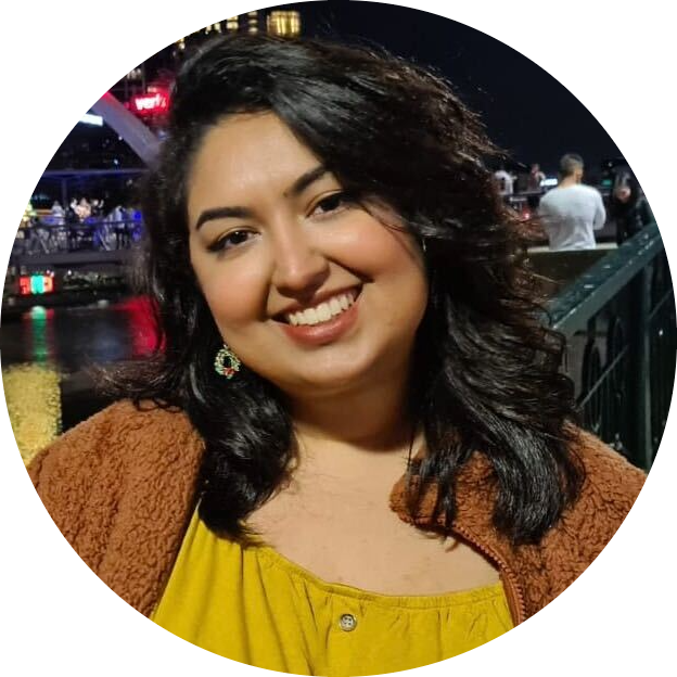 An image of our current president Tiara Pires - she's facing the camera smiling with her head tilted to the side. She's wearing a yellow top and a light brown cardigan, and appears to be standing outside in a nighttime city environment.