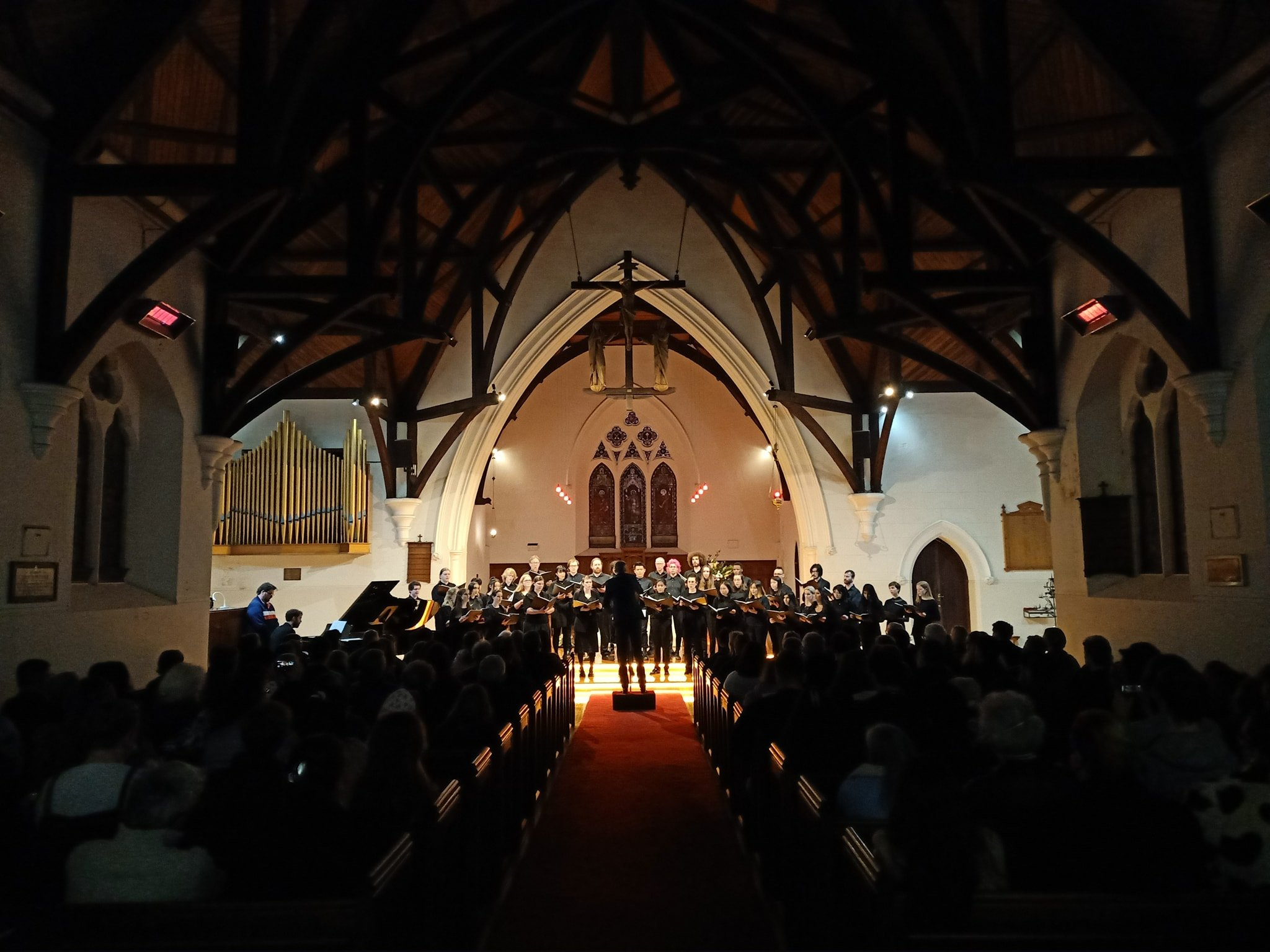 A picture of the packed insides of St John's East Malvern as we were performing our concert! There are packed pews, an empty red-carpeted walkway in the middle, and the choir on stage lit up and holding our music folders as we stand and sing.