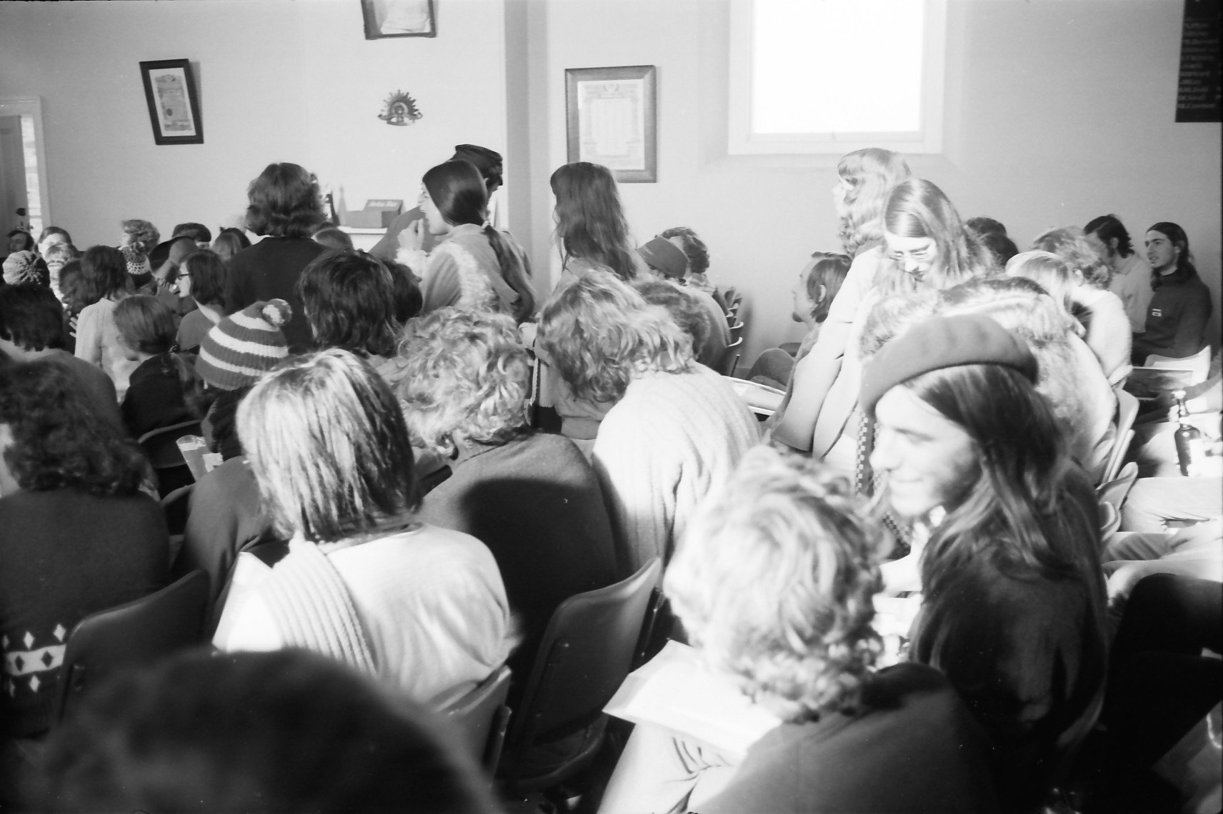 A black-and-white image of a previous camp rehearsal! We see people sitting in rows of chairs that are diagonal across the image. Near the front, somebody is smiling at their neighbour and likely chatting, but most people appear to be looking forwards. The walls of the room are a light colour, there are a few frames hanging on the wall, and we see the room's window in the top-right quadrant of the image.