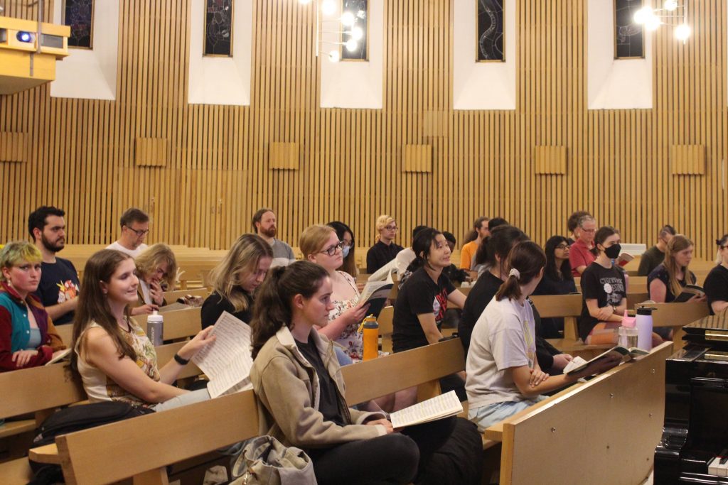 An image of a rehearsal in flow! We see the pews within the Main Chapel of the Religious Centre where we rehearse, at a diagonal angle across the bottom third of the image. They're a light brown colour, and many choristers sit on them while holding their sheet music books and looking in a variety of directions. The back walls are a similar light brown, and panelled in thin vertical slits. We also see a number of dark stain-glass  thin rectangular windows, set in larger white rectangular panels amidst the brown walls. In the bottom-right corner we see a smidgeon of the piano, and in the top-left a little bit of a balcony that is in the room.