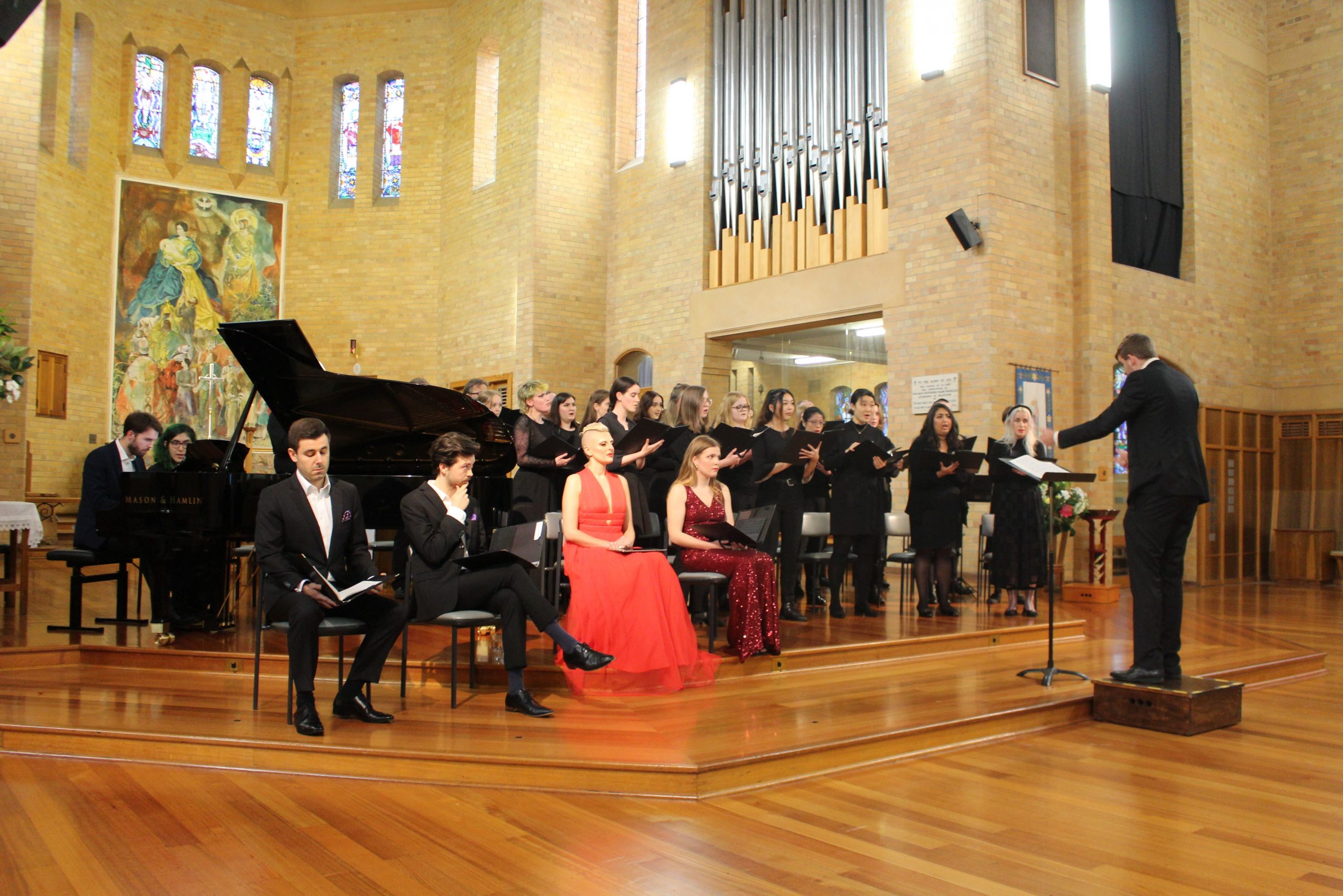 Our choir mid-performance of Stabat Mater! You can see our four soloists seated in chairs near the front of the stage on the left of the image, in front of the piano (behind which two figures sit, one playing and one page-turning). Our conductor Lachie busily conducts, standing on the right of the image in front of a music stand, and we see the choir standing in rows busily singing next to the piano and behind the soloists. The environment is light brown and churchy - we see what looks like a pipe organ and an intricate painting in the background.