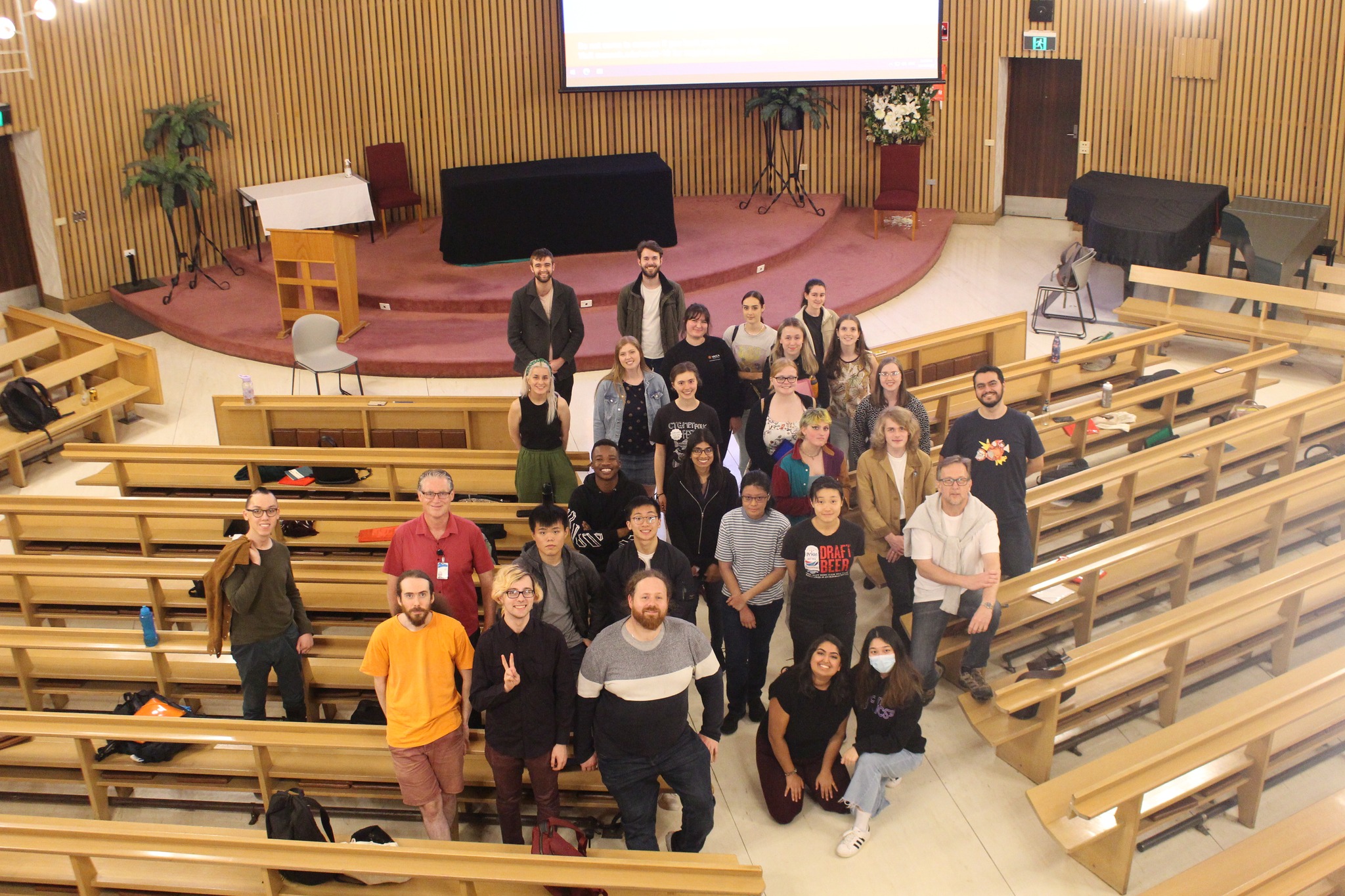 A high-angle picture taken in the Religious Centre of our choir! Most of the 30ish choristers are standing amidst the space between the light brown pews (though a couple of people are kneeling on the floor) and mostly everyone is smiling towards the camera.