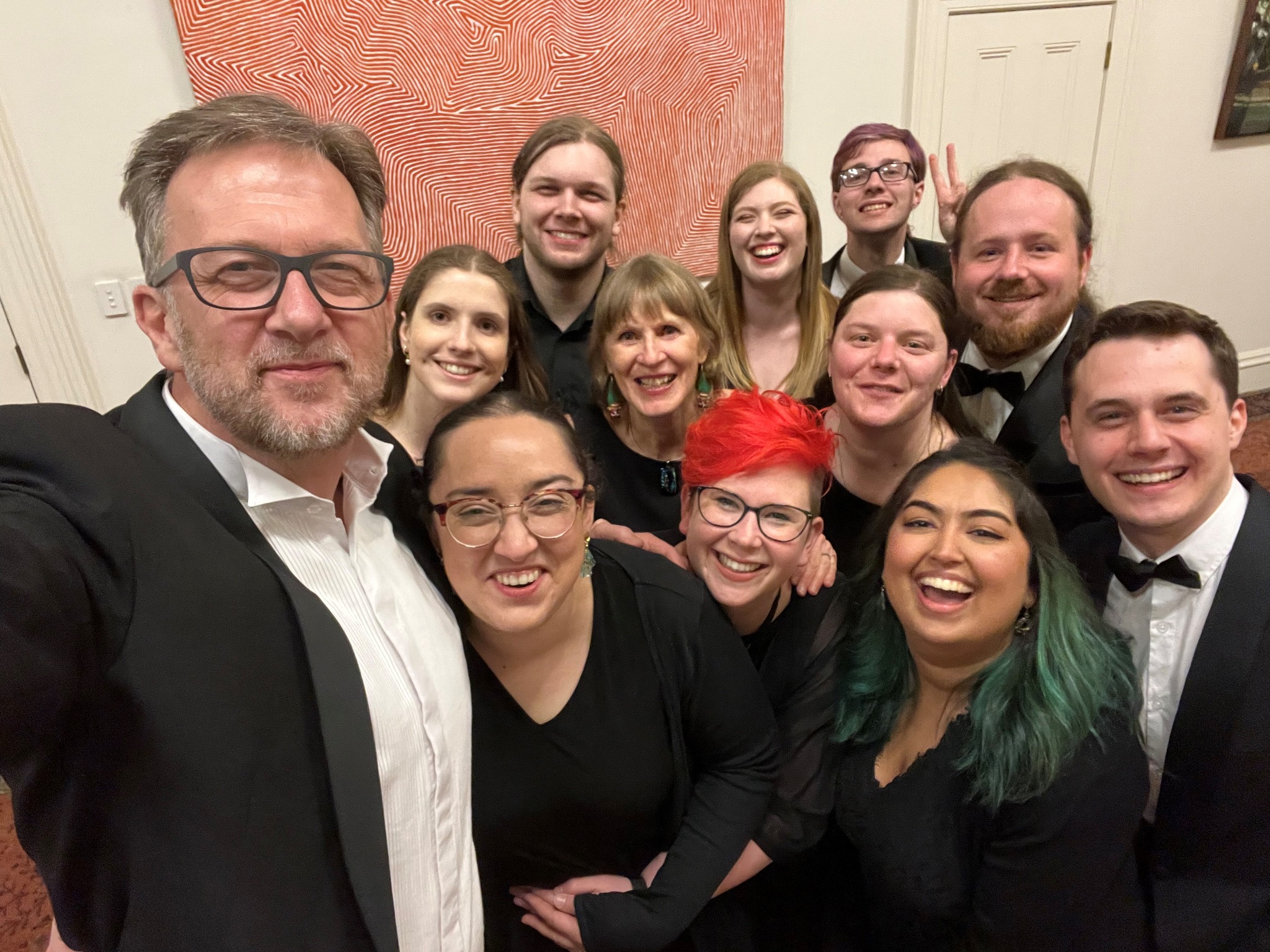 A selfie of a group of choristers after a carolling gig! Everyone is dressed very smartly - black suits and white shirts, dresses, ties, the like - and looking towards the camera and smiling. In the background we see the cream walls of the room and a painting made of interesting red curve shapes.