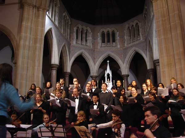 A MonUCS concert in progress! We see the conductor towards the left of the image, facing towards and conducting a group of choristers. They all stand wearing formal black clothing and holding sheet music, while some string players sit in front of them playing their instruments. The setting appears to be a cathedral - we see a round space in grey-brown stone with many archways. 