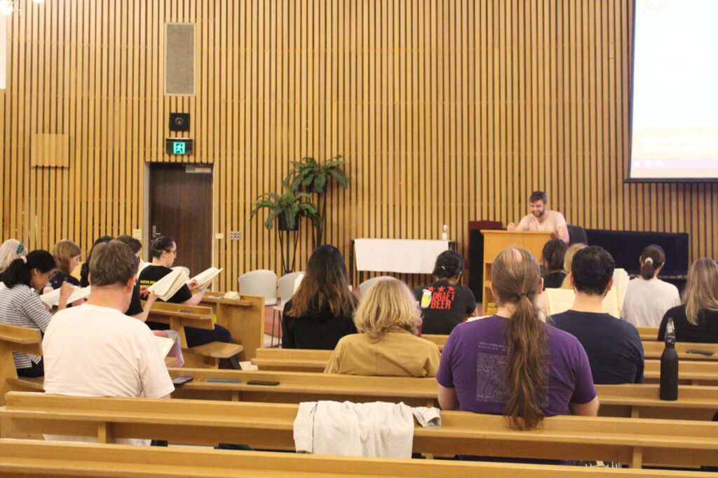 A fairly standard rehearsal in the Religious Centre. From the perspective of the pews, we see a number of choristers sitting in rows, looking at their music as our conductor Lachie sits at the front facing everybody. 