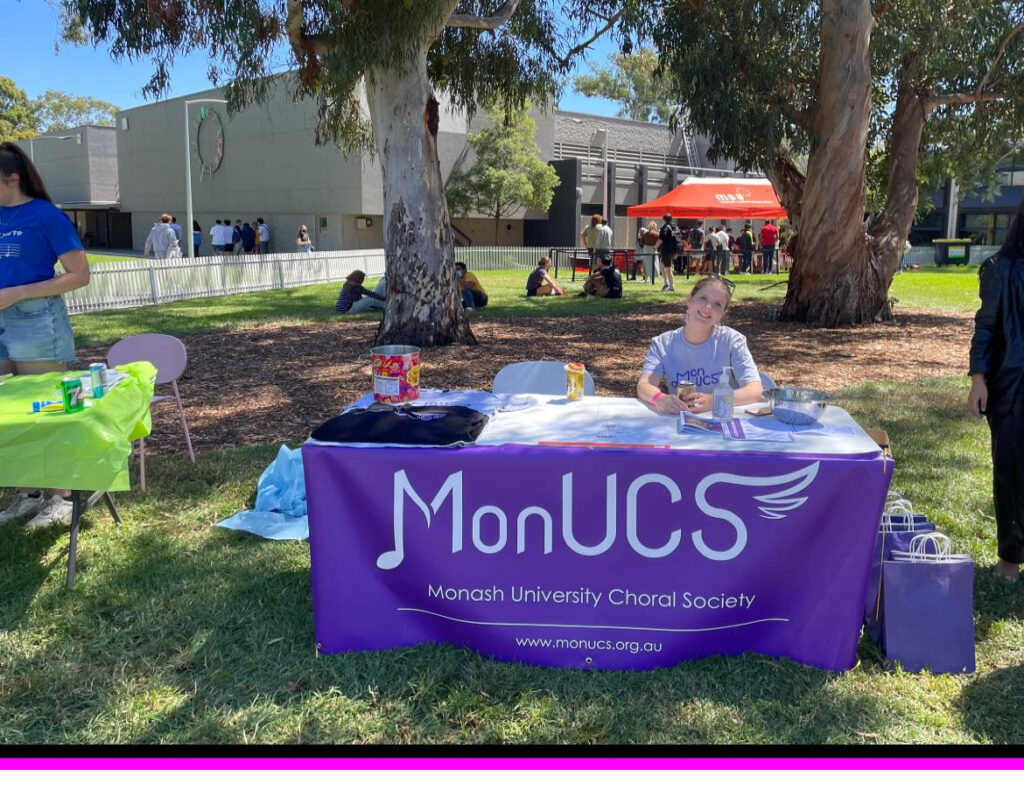 A picture of our O-Week stall from last year! It's a table with a purple MonUCS banner hanging from the front of it, in an outside grassy environment on Clayton campus. Our secretary Anna Tinney is sitting at a seat at the table and smiling. You can also see some purple goodie bags to the right of the image.