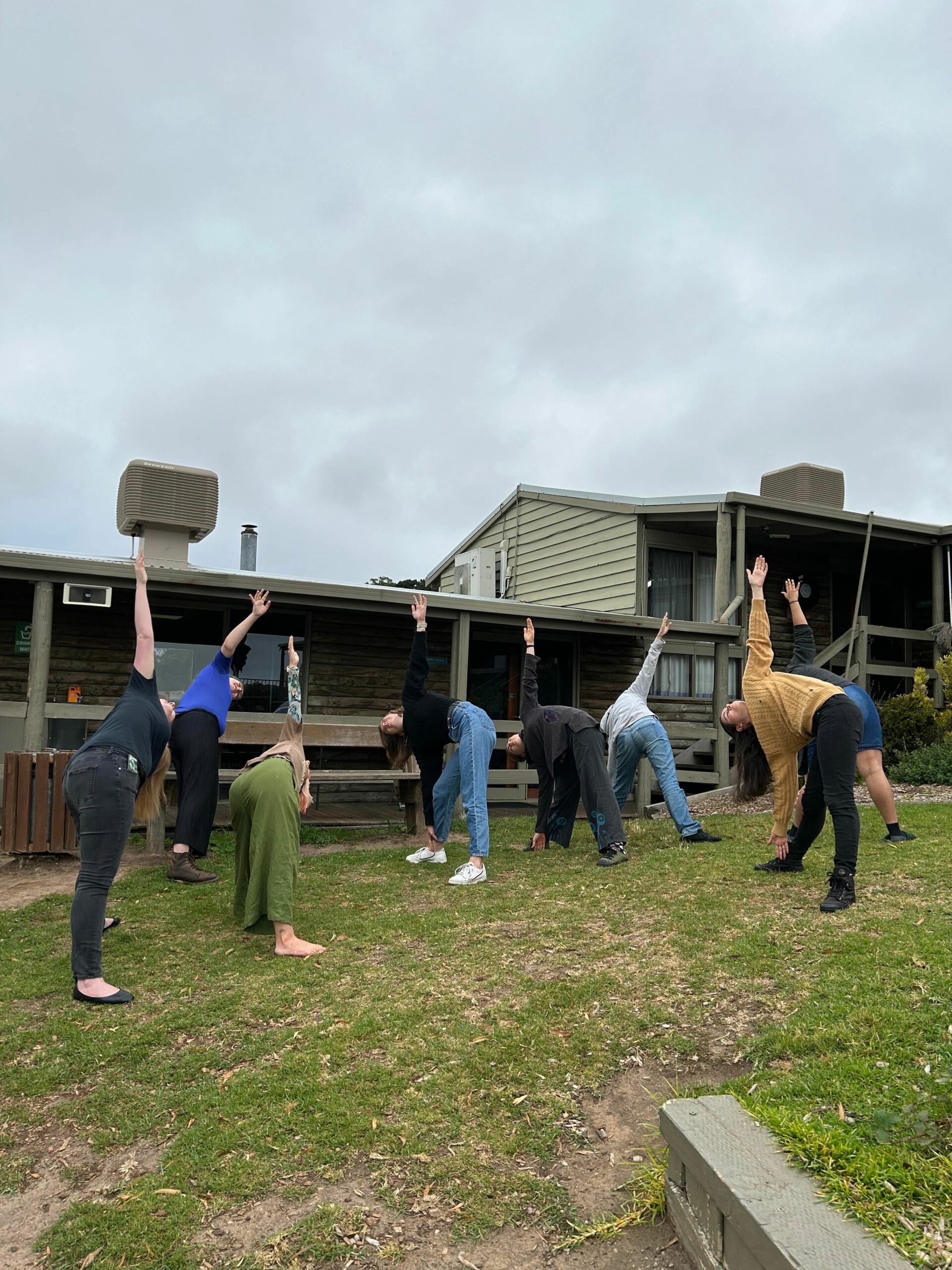 Outside the campsite buildings on a hilly grassy area, a number of choristers are bent down with one hand in the air, engaged in yoga