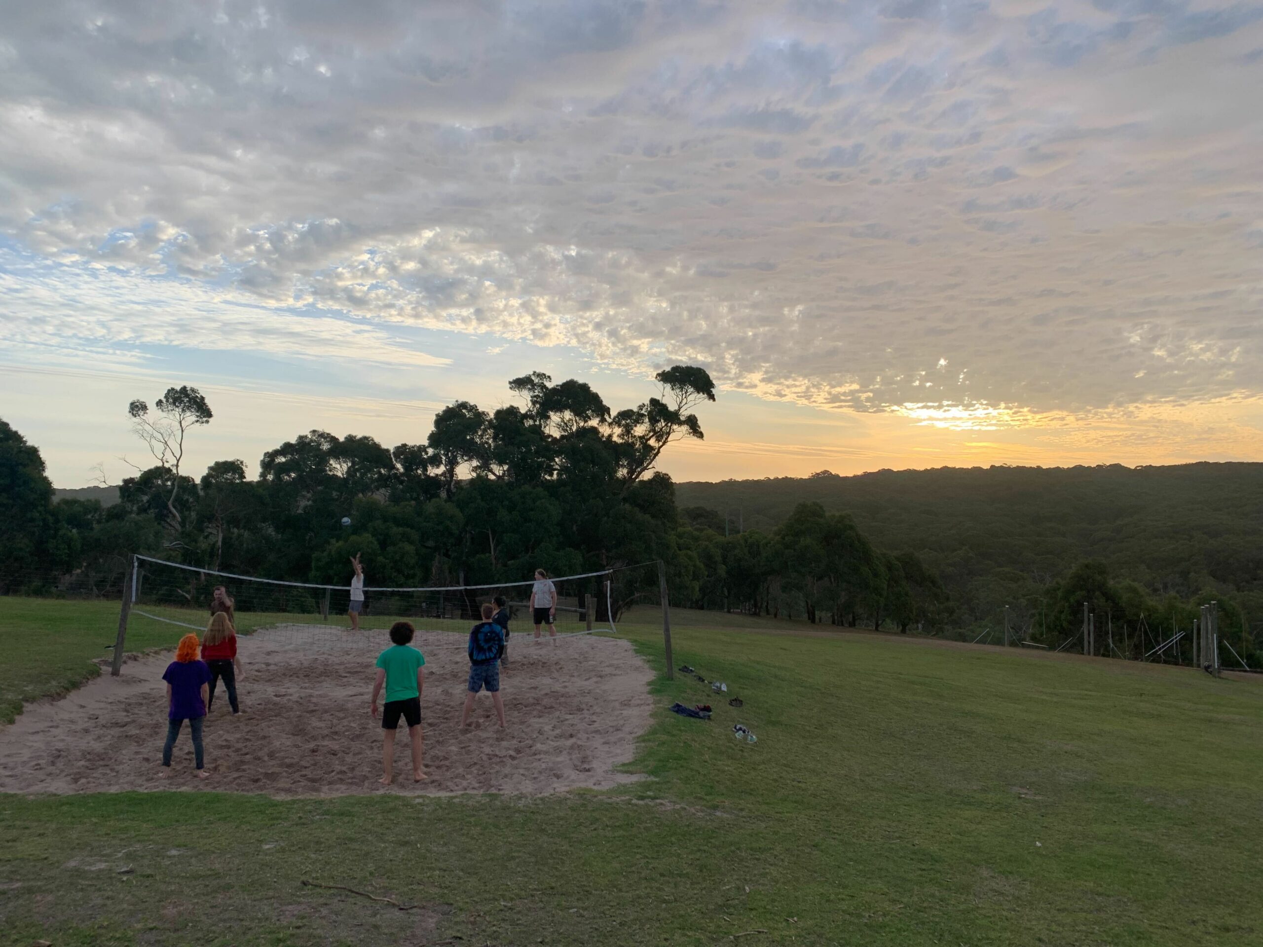 Outside in early evening (setting sun, cloudy sky) a small sandy volleyball pitch sits within a larger grassy area, and some choristers stand on either side playing