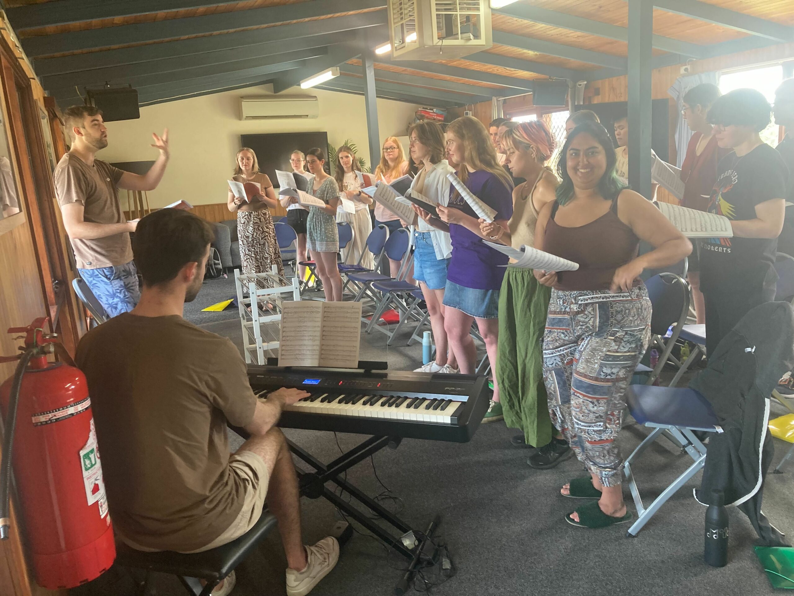 Another busy moment of rehearsal - our perspective is from behind our pianist, and we see standing rows of choristers holding sheet music, a couple of whom look towards the camera and smile