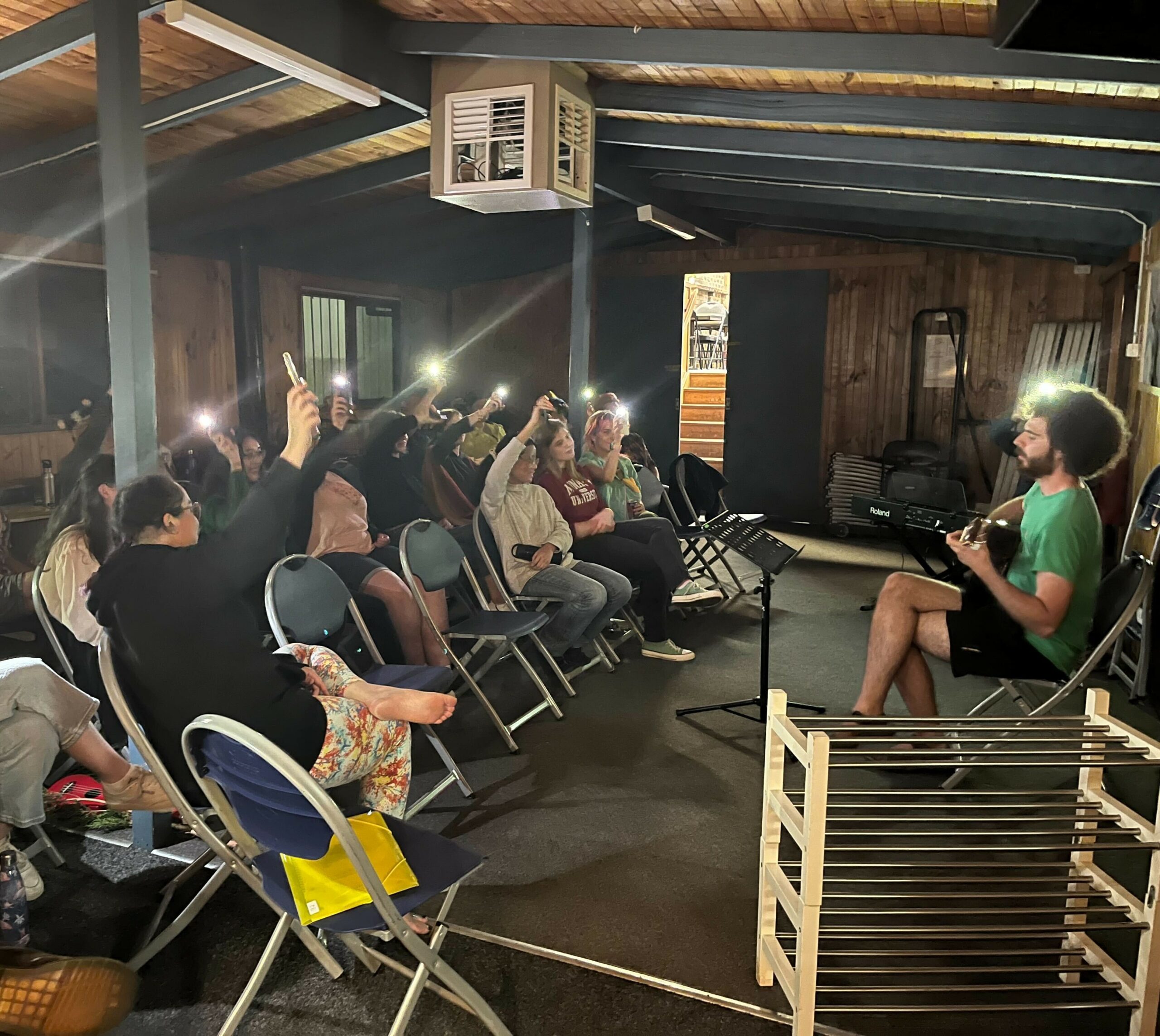 A moment from our Revue - a guitarist plays and sings sitting on a chair in the right of the image, while towards the left rows of people sing along while waving phones with lit torches