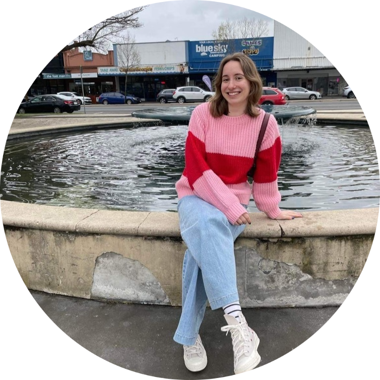 Grace is sitting on what looks like the edge of a large fountain with her legs crossed, wearing a pink and red jumper and light blue jeans, smiling into the camera