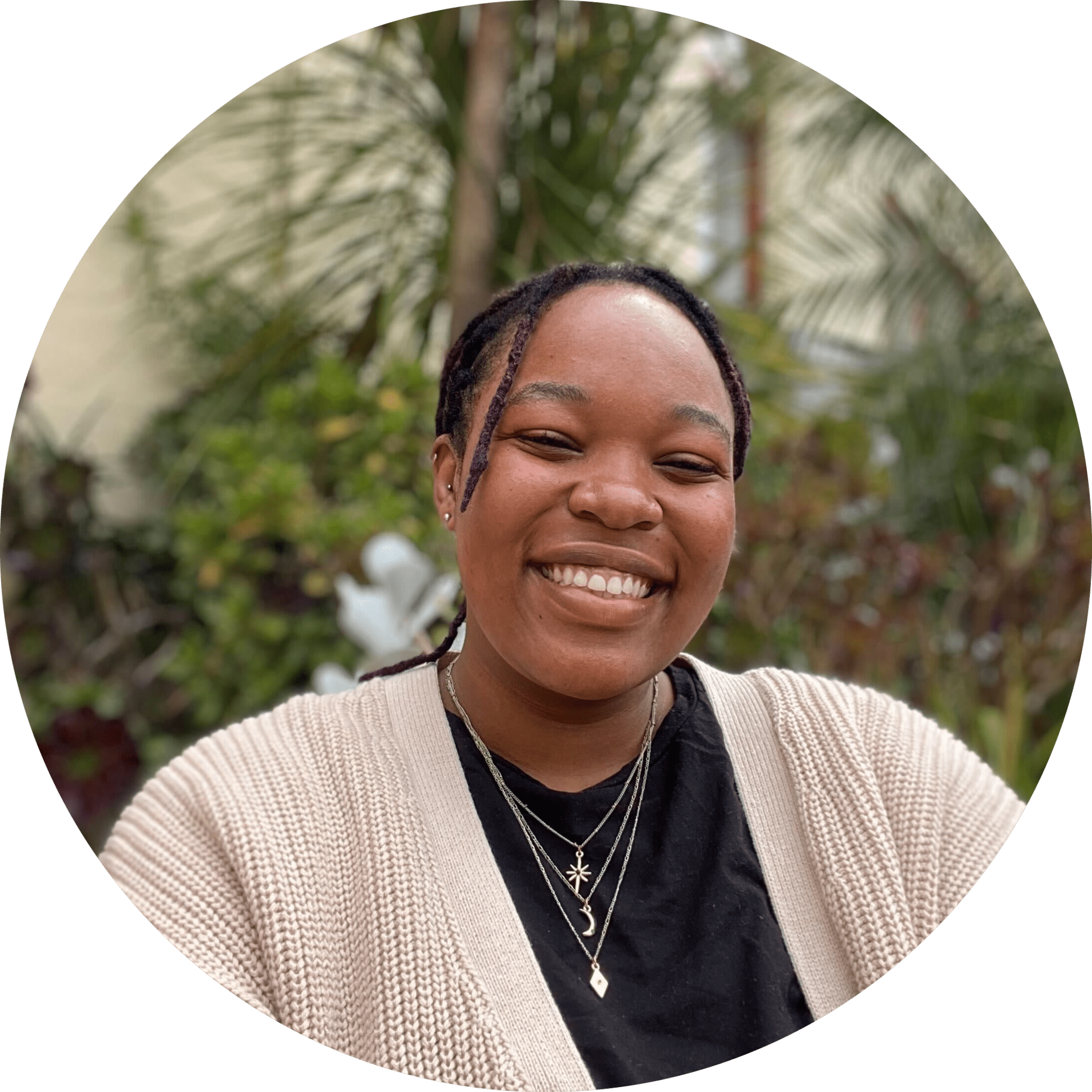 Sibu smiles into the camera in front of a leafy green backgroudn. THey are wearing a light cream cardigan over a black t-shirt with a number of silver pendant necklaces on