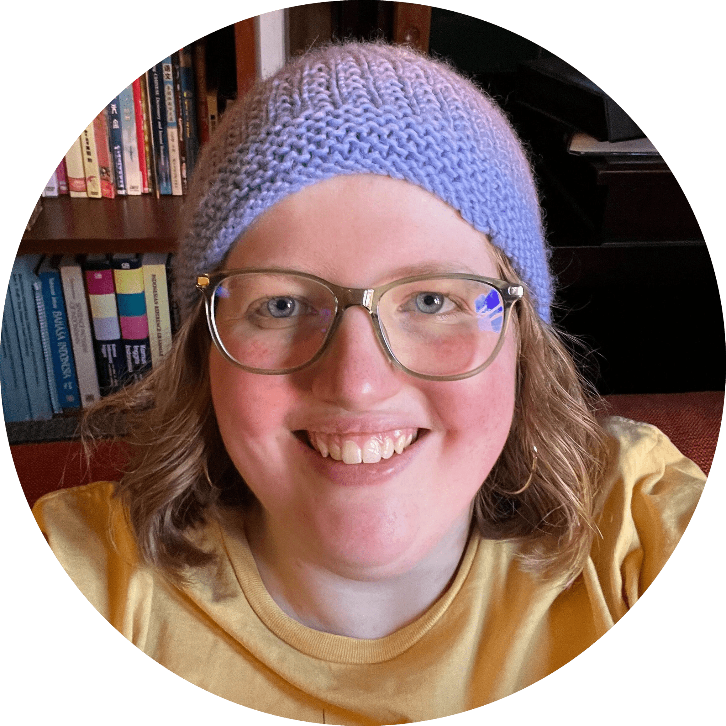 Jas smiles into the camera, wearing glasses, a light-blue beanie and a yellow shirt, in front of a homely background with filled bookcases and shelves